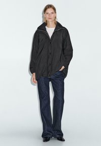 Svart lättviktsjacka med hög krage, knappstängning och långa ärmar, ihop med mörkblå jeans och svarta skor.