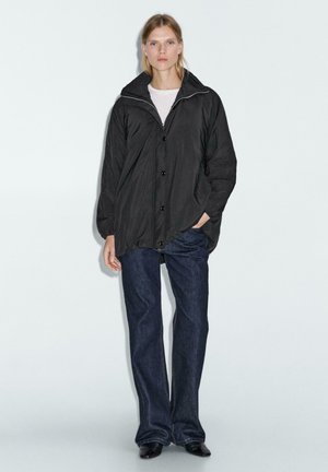 Giacca nera leggera con colletto alto, chiusura con bottoni e maniche lunghe, abbinata a jeans blu scuro e scarpe nere.