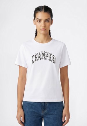 Junge Frau mit geflochtenem Haar, die ein weißes T-Shirt mit dem Schriftzug "CHAMPION" in schwarzen gemusterten Buchstaben und eine blaue Jeans trägt, vor einem weißen Hintergrund steht.