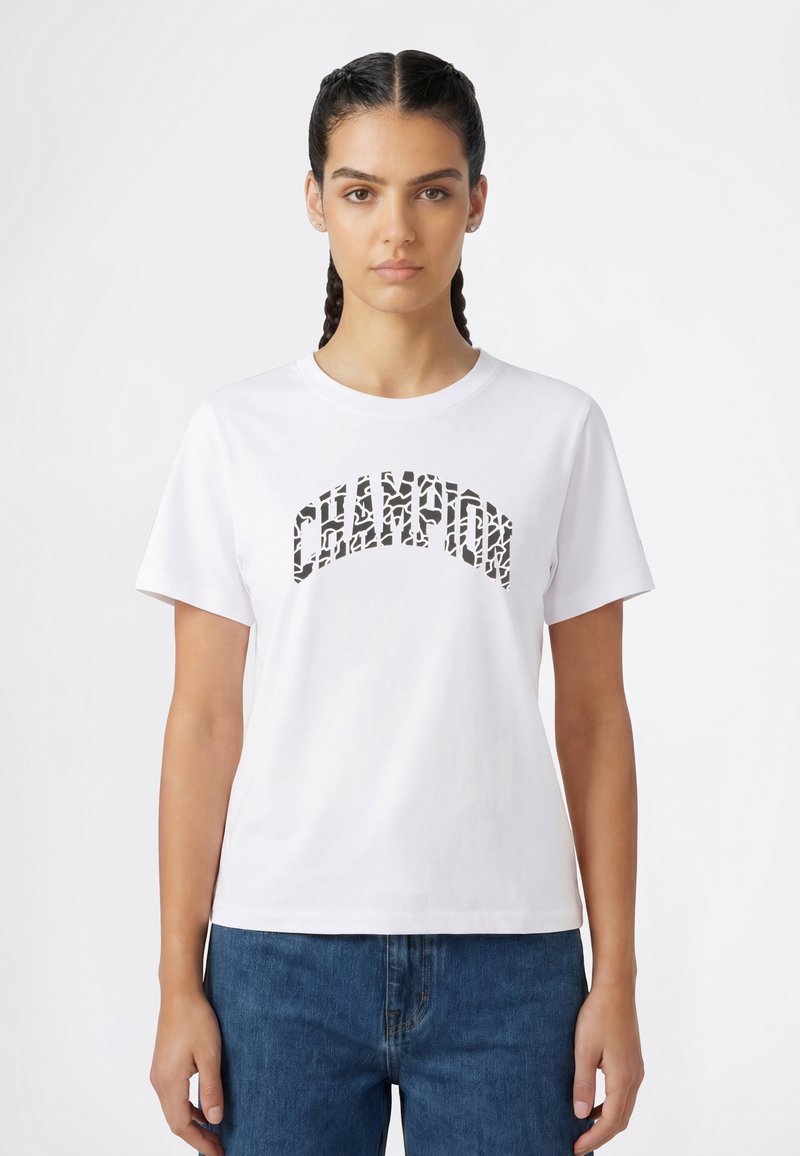 Jonge vrouw met gevlochten haar, gekleed in een wit T-shirt met "CHAMPION" in zwarte patroonletters en een blauwe spijkerbroek, staand tegen een witte achtergrond.