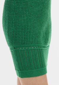 Grøn strikket sweaterdetalje med åben vævet tekstur, ribbet kant og horisontale striber med små udskæringer nær manchetterne.