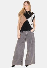 Top senza maniche lavorato a maglia con motivi geometrici in nero, marrone, bianco e accenti di rosso; abbinato a pantaloni di jeans grigi a gamba larga.