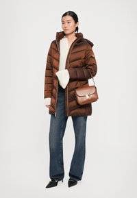 Vrouw in een bruine pufferjack, crème trui, blauwe jeans, zwarte puntige hakken en met een bruine leren handtas aan haar arm.