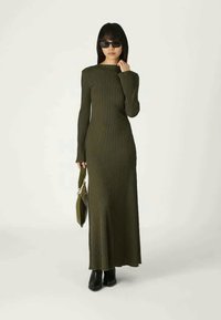 Γυναίκα σε μακριά, ελαιόχρωμη ριγέ φόρεμα με μακριά μανίκια, μαύρες μπότες, ηλιακά γυαλιά, κρατώντας μια ταιριαστή ελαιόχρωμη τσάντα χεριού.