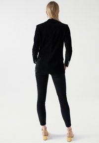 Černý sartorialní blazer s dlouhými rukávy, zkombinovaný s přiléhavými černými kalhotami a sandály na zlatém podpatku. Hladká textura a moderní design.