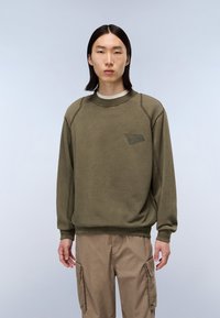 Oliven grøn sweatshirt med rund hals og ribbet kant, med en diskret logo detalje på brystet. Blød tekstur, afslappet pasform.