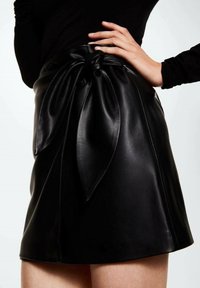 Femme portant une jupe en cuir noire avec un grand nœud noué à la taille, associée à un haut noir à manches longues.