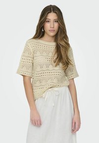 Top de ganchillo en beige claro con mangas cortas, que presenta un patrón de calado y un escote redondo, combinado con una falda blanca con cordón.