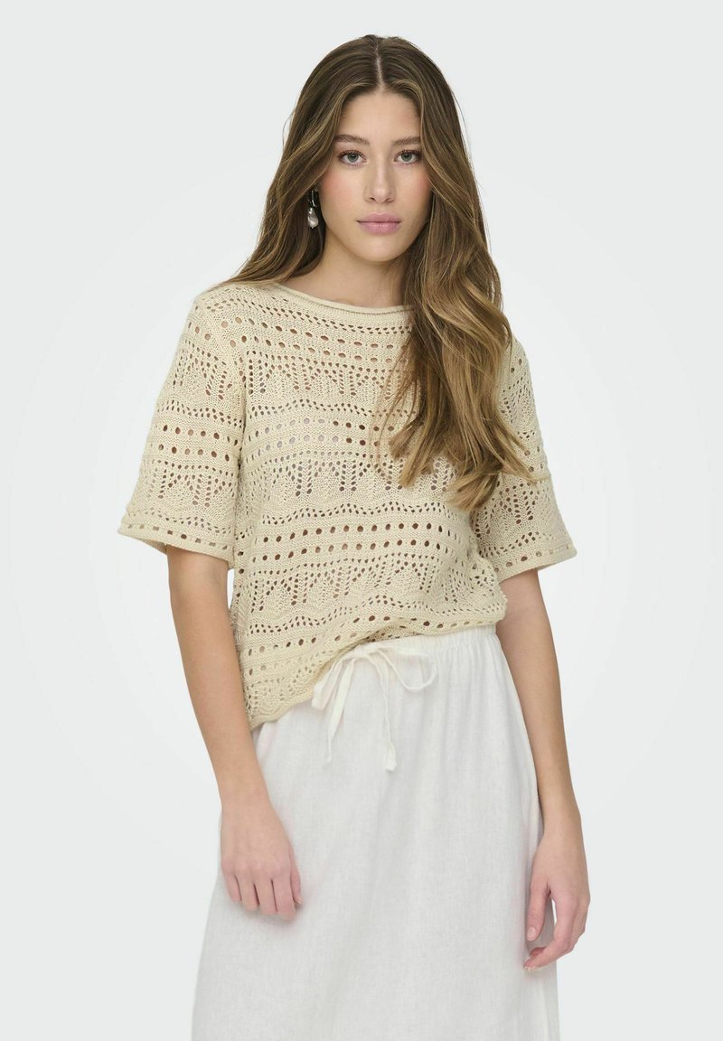 Top de ganchillo en beige claro con mangas cortas, que presenta un patrón de calado y un escote redondo, combinado con una falda blanca con cordón.