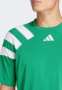 Roheline sportlik särk valgete õlavarre triipudega ja ümar kaelus. Esiküljel on nähtav logo vasakus ülanurgas ning särk on valmistatud siledast kangast.
