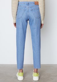 Svetle modré džíny z denimu s vysokým pasem, rovnými nohavicemi a dvěma zadními kapsami. Nositel má na sobě bílé tenisky s neonově zelenými akcenty.