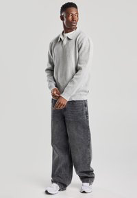 Harmaa neule pitkähihainen pusero, jossa on kauluspää, yhdistettynä löysiin mustiin farkkuliiveihin. Mallilla on päällään valkoiset lenkkarit. Yksinkertainen tausta.