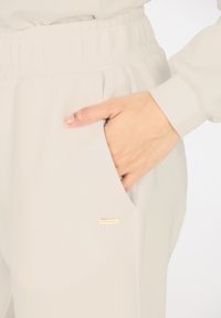 Helle beige Hose mit einem elastischen Bund. Sie verfügt über eine Seitentasche und ein kleines goldenes Akzentlabel. Glatte, weiche Stofftextur.