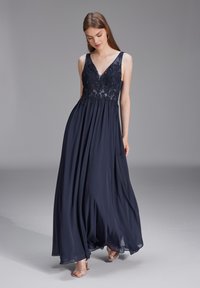 Robe de soirée bleu marine avec un décolleté en V profond, un corsage en dentelle orné et une jupe fluide qui atteint le sol. Matière en chiffon lisse.