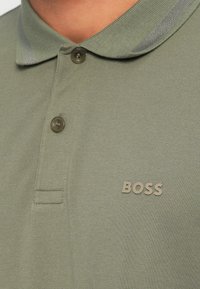 Oliwkowa koszulka polo z fakturowanej tkaniny, z dwukolorową plisą zapinaną na dwa guziki oraz małym, wypukłym logo "BOSS" na piersi.