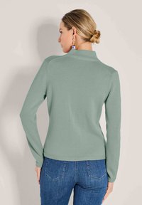 Frau mit blonden Haaren in einem Dutt trägt einen hellgrünen Pullover und bluejeans, steht mit dem Rücken zu einem schlichten hellen Hintergrund.