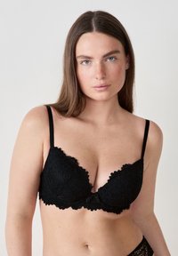 Schwarzer Spitzen-BH mit gewellten Kanten, tiefem Ausschnitt, verstellbaren Trägern und einem kleinen Schleifchen in der Mitte. Weicher, strukturierter Stoff.