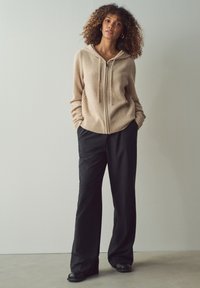 Felpa beige con zip, bordo e polsini a coste, abbinata a pantaloni neri a gamba larga. Il modello è in piedi con le mani nelle tasche su uno sfondo neutro.