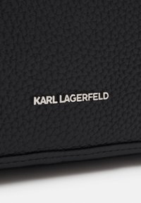 Pelle nera testurizzata con una forma arrotondata, caratterizzata dal logo in rilievo "KARL LAGERFELD" in argento, bordi lisci e dettagli di cucitura visibili.