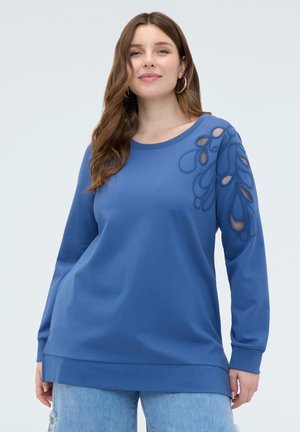 Donna con top a maniche lunghe blu con ricamo floreale traforato sulla spalla, abbinato a jeans chiari strappati, in piedi contro uno sfondo chiaro.