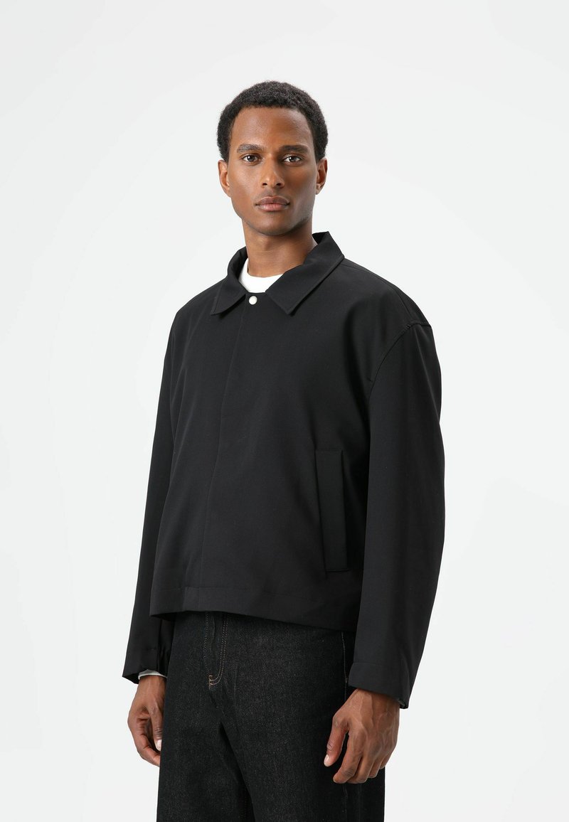 Black cropped jacket with a pointed collar, featuring a front button closure and side pockets. Smooth fabric with a minimalistic design.