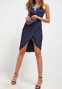 Žena v námořnickém modrém bezramínkovém šatu se stříbrnou výšivkou na v ýstřihu, držící černou kabelku a v černých sandálech na kotníky.