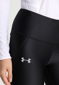 Černé sportovní legíny vyrobené z elastického materiálu. Mají vysoký pas a malé šedé logo na dolní části levé nohy. Hladká textura.