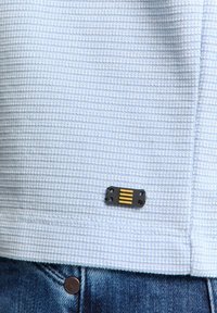 Lichtblauw, gestructureerd stoffen overhemd met een zwart en geel hardware accentlabel vlakbij de zoom, gecombineerd met blauwe jeans.