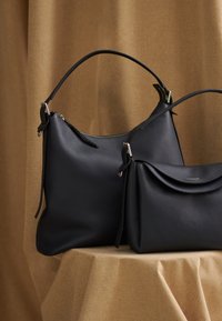 To sorte tasker i læder placeret på en tekstureret overflade. Den ene taske har en løs form med skulderrem, den anden er struktureret med en klap.