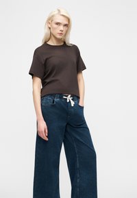 Ξανθιά γυναίκα που φοράει σκούρο καφέ κοντομάνικο πουκάμισο και πλατύχωρα μπλε τζιν παντελόνια με λευκό κορδόνι, στέκεται με το ένα χέρι στην τσέπη.