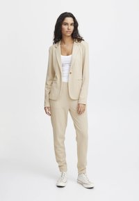 Ljust beige skräddarsydd kostym med en blazermodell med en knapp och matchande joggingbyxor, med subtila vertikala ränder, bärs över en vit topp.