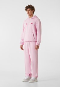 Svetlo roza kapuca z vezenimi detajli, kenguru žepom in ujemajočimi se trenirkami. Outfit se ponaša z udobnim krojem in mehkobo materiala.