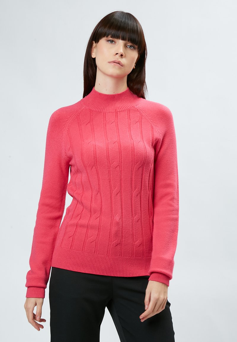 Rosa Strickpullover mit hohem Ausschnitt, strukturiertem Kabeldesign und gerippten Bündchen und Saum. Das Model trägt schwarze Hosen.