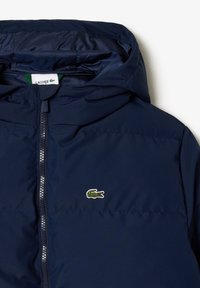Doudoune marine avec capuche, dotée d'une fermeture éclair à l'avant et d'un petit logo crocodile vert sur la poitrine. Texture de tissu lisse et brillante.
