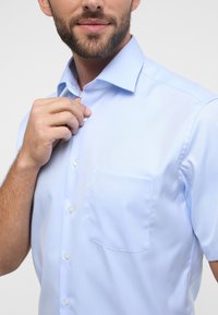 Camicia azzurro chiaro a maniche corte con colletto, bottoni frontali e una piccola tasca sul petto. Tessuto morbido con un design pulito.