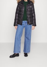 Schwarze Steppjacke mit pelzgefütterter Kapuze, Reißverschluss vorne, gesteppte Musterung; grüner Strickpullover darunter; hoch taillierte blaue Jeans mit weitem Bein; schwarze Schuhe.