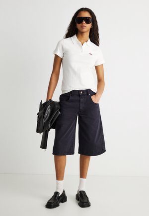 Woman wearing black sunglasses, white polo shirt, black wide-leg knee-length shorts, white socks, black loafers, holding black leather jacket.