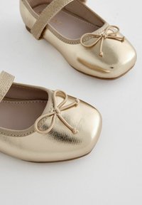 Ballerines dorées avec une finition métallique brillante, bout rond, un nœud décoratif et une sangle élastique sur le coup de pied. Texture lisse.