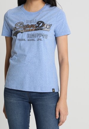 Kvinde iført lyseblå SuperDry t-shirt med sort og sølv logo og mørkeblå jeans, stående mod en ensfarvet baggrund.