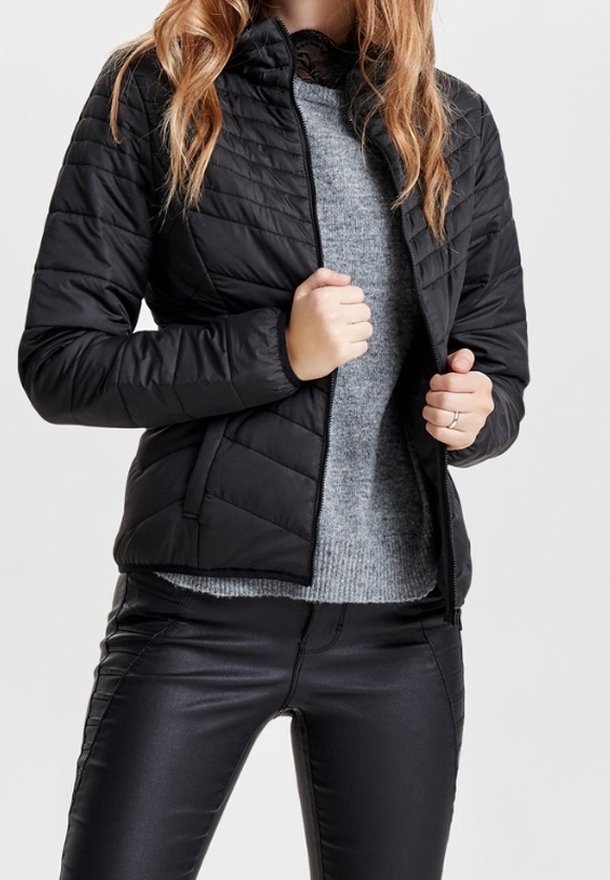 Czarna pikowana kurtka z wysokim kołnierzem, zamkiem z przodu i teksturowanym materiałem. Noszona na szarym swetrze i czarnych spodniach, podkreślająca warstwowy wygląd.