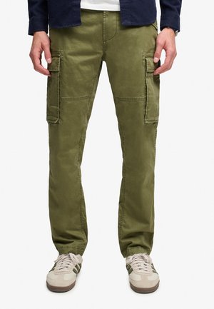 Mann trägt olivgrüne Cargohosen mit großen Seitentaschen und beige Sneaker mit grünen Streifen, steht vor weißem Hintergrund.