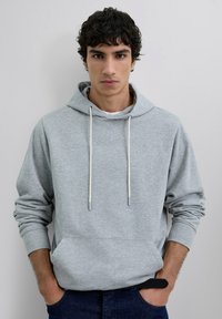 Sudadera de algodón gris claro con un bolsillo frontal, capucha con cordón y puños acanalados. Llevada sobre una camisa blanca y combinada con jeans oscuros.