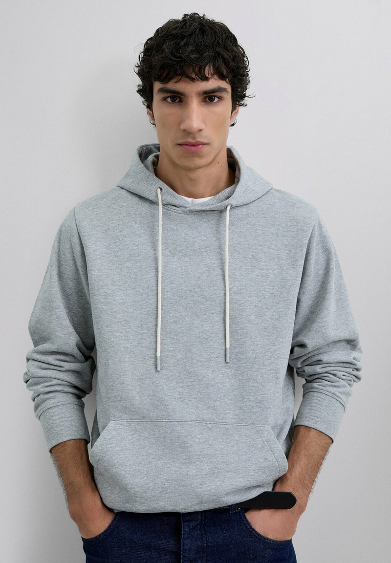 Sudadera de algodón gris claro con un bolsillo frontal, capucha con cordón y puños acanalados. Llevada sobre una camisa blanca y combinada con jeans oscuros.