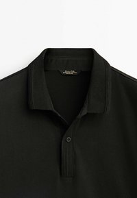 Polo noir avec une texture douce en coton, doté d'un col classique, d'une patte à deux boutons et de subtils accents côtelés au niveau du col.
