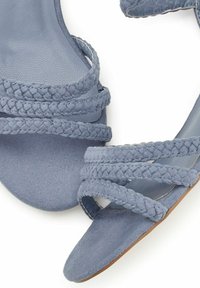 Blå flade sandaler med fletstropper, suede tekstur, åben tå design og en glat indersål. Sålen har en lys brun farve.