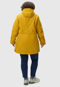 Yellow waterproof parka with a hood, featuring a straight design, snap-button cuffs, and a smooth fabric texture. Worn over dark jeans.