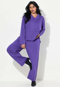 Lila Loungewear-Set bestehend aus einem langärmeligen Oberteil mit V-Ausschnitt und lockerer Passform, kombiniert mit weit geschnittenen Hosen. Weicher, glatter Stoff.