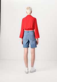 Czerwona krótka bluzka z dzwonkowymi rękawami, połączona z jasnoniebieskimi szortami dżinsowymi i białymi trampkami za kostkę. Jednolity tle, widok z tyłu.