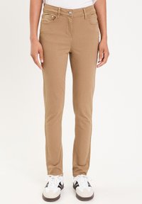 Pantalons en toile beige avec une coupe ajustée, poches avant, fermeture à bouton en métal et détails de couture minimalistes, portés avec des chaussures sportives blanches et noires.