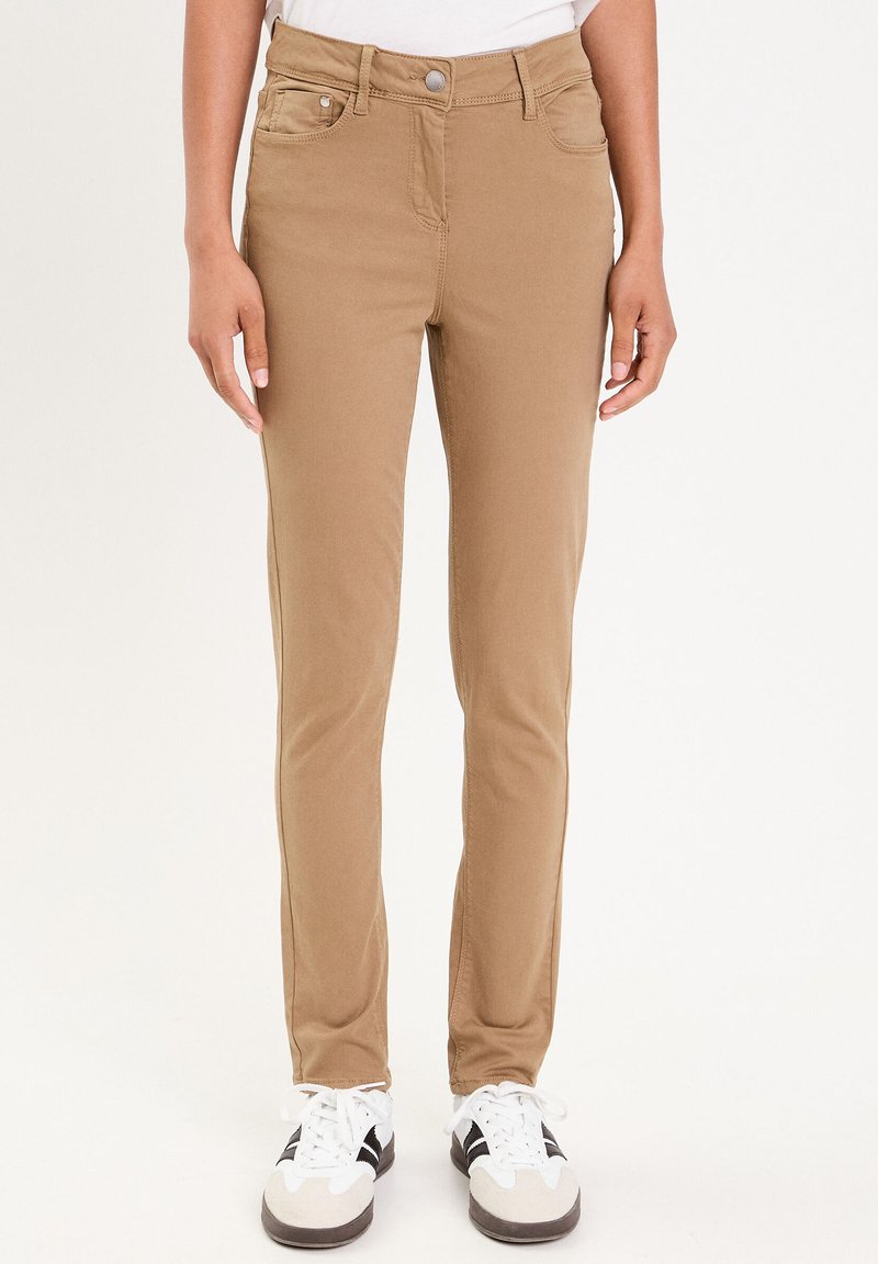 Beige Baumwollmischungshose mit schmaler Passform, vorderen Taschen, Metallknopfverschluss und minimalen Steppdetails, getragen mit sportlichen schwarz-weißen Schuhen.